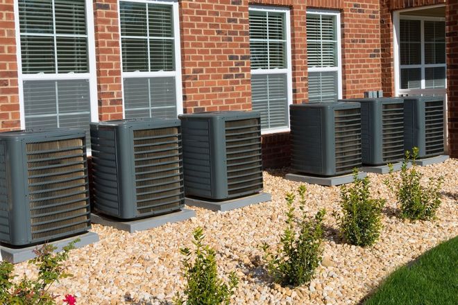 Row of outdoor air conditioning units beside a brick building with windows and shrubs