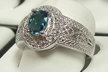 A silver ring with a large oval teal gemstone and a halo of small white stones. The band features intricate filigree detailing.