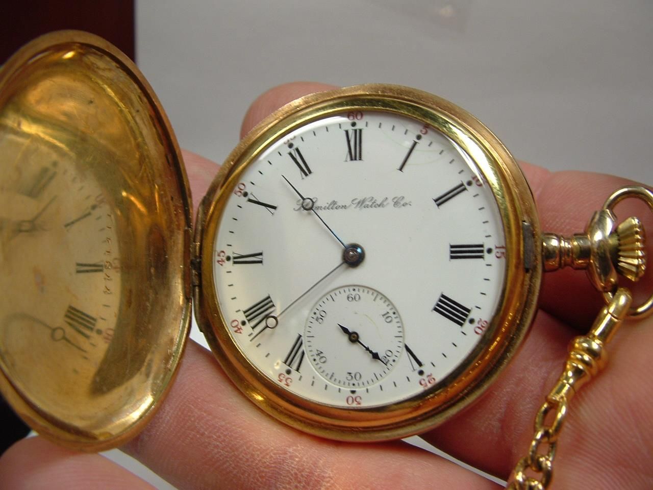 Gold pocket watch with a white face, Roman numeral hour markers, and a second dial. The open case is visible, held by a hand with a gold chain attached.