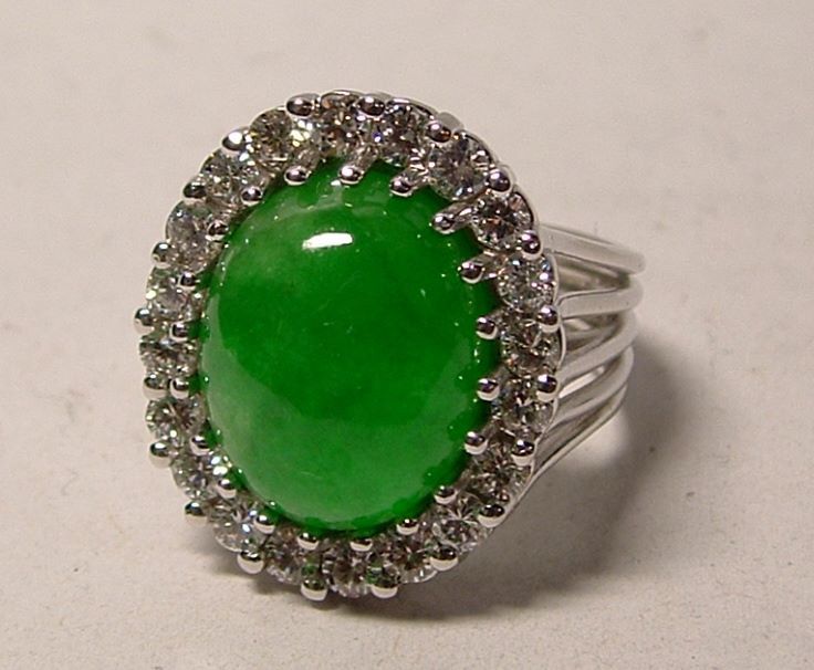 Oval, bright green jade ring with a halo of white gemstones, set in a silver band with multiple prongs.