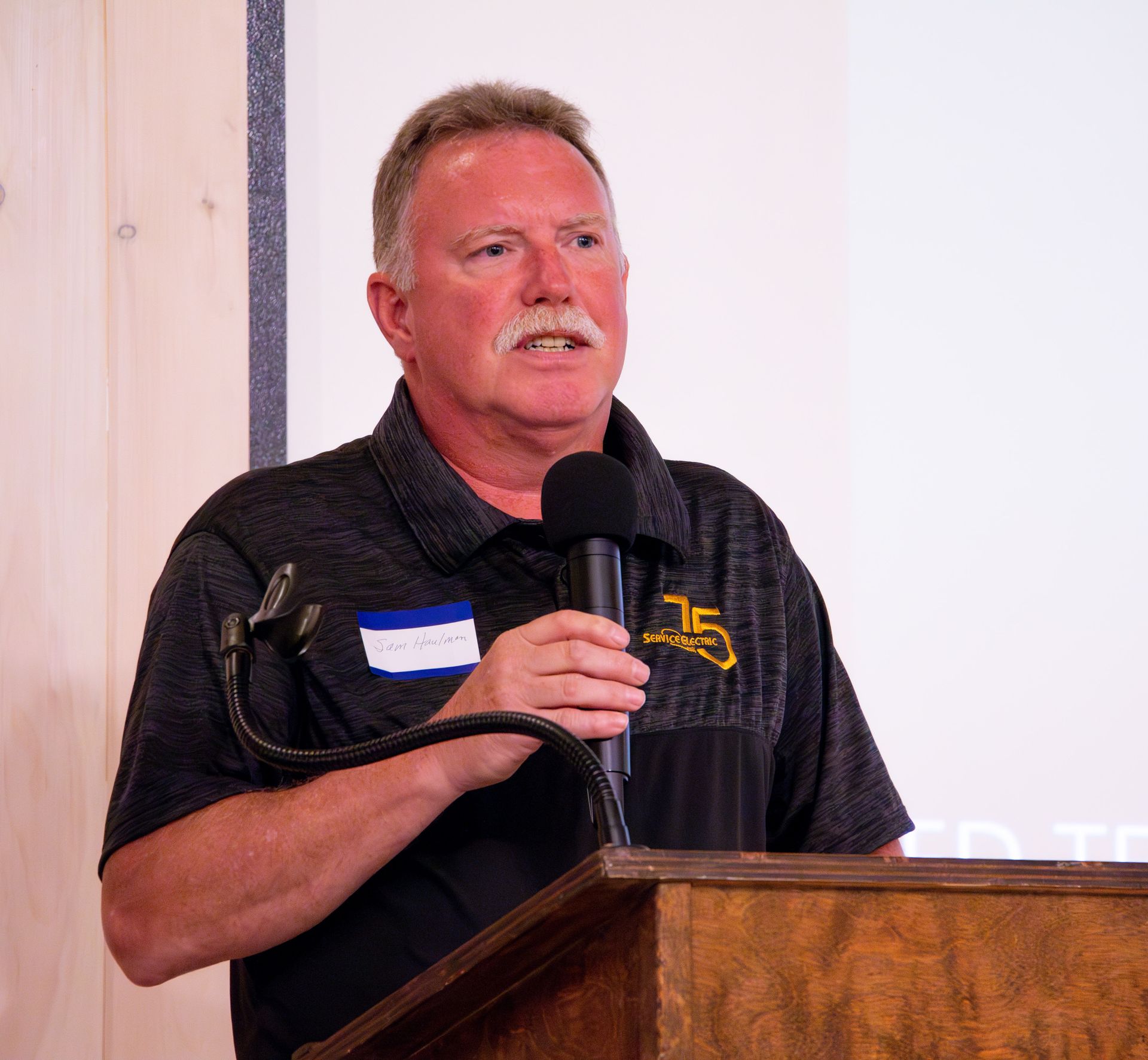 A man with a mustache is standing at a podium holding a microphone.