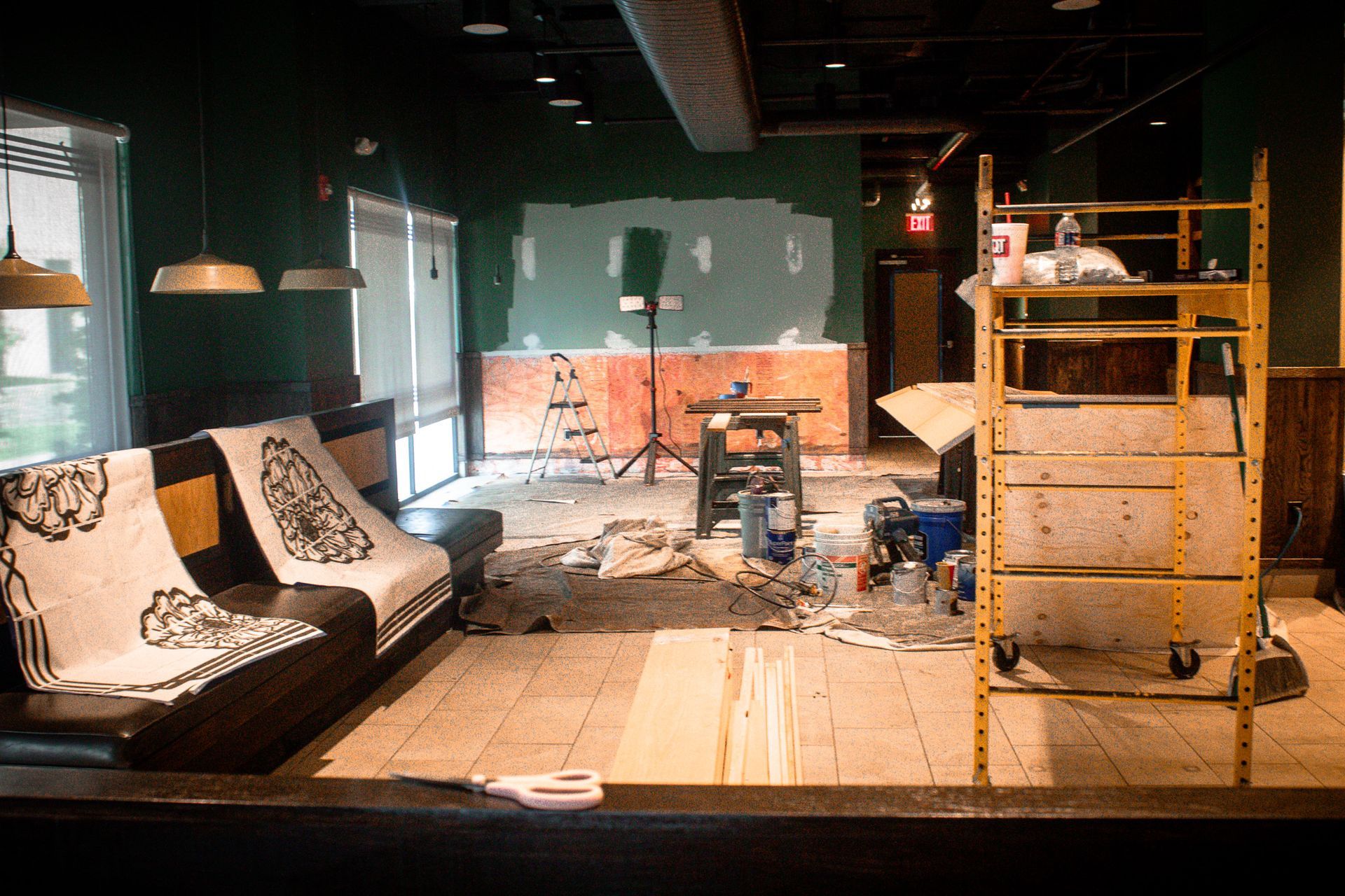A cluttered living room with a sofa, wooden floor, ceiling lights, and a partially built shelving unit.