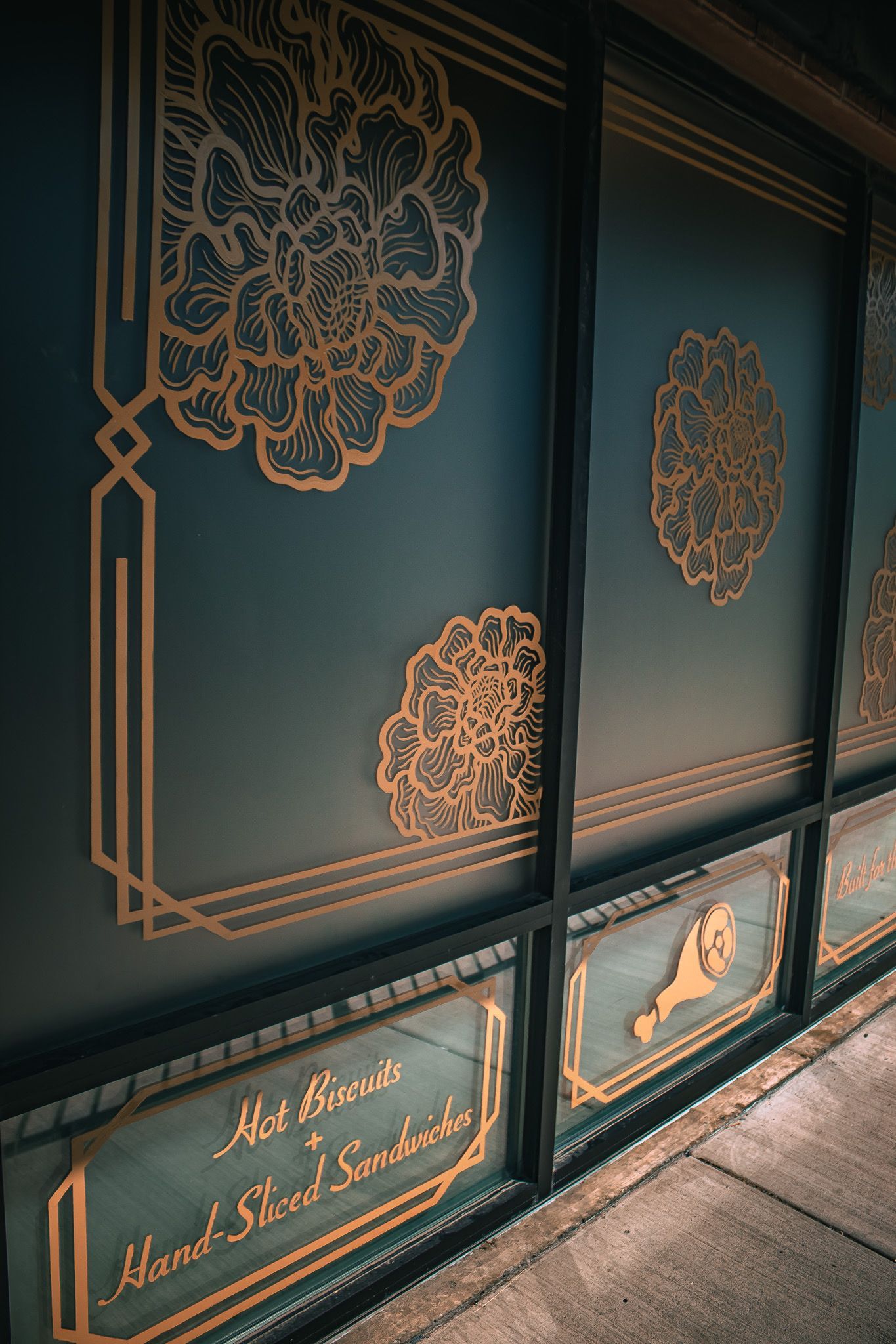 Teal decorative storefront panels with gold floral line art and framed trim beside a wooden floor.