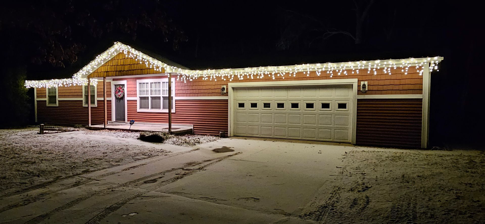 Snow-covered house at night, lit with Christmas lights and a two-car garage.