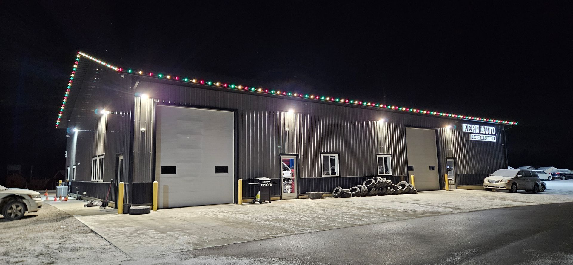 Small industrial building at night with holiday lights, parked vehicles, and snowy ground.