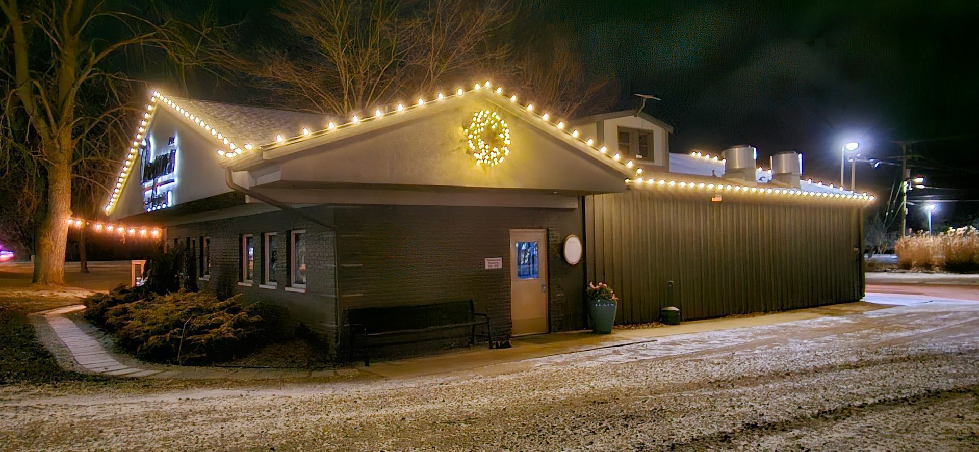 Small building lit by string lights at night, with snow on the ground and trees in the background