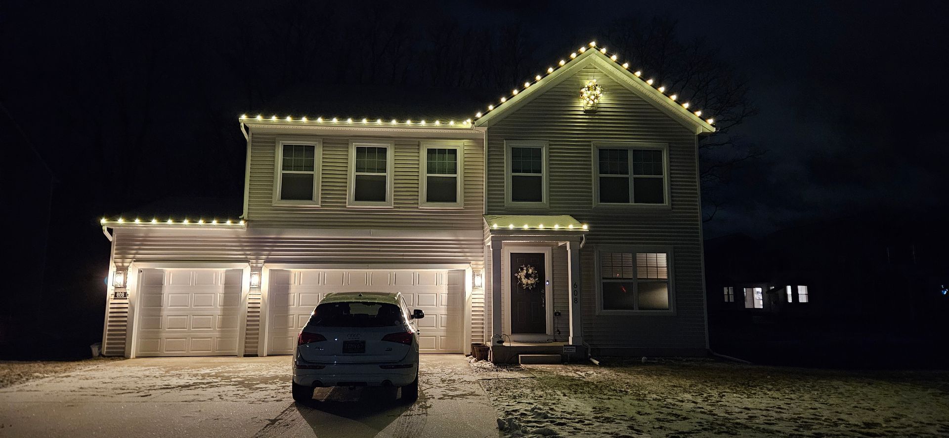Snowy suburban house decorated with white Christmas lights, with a car parked in the driveway at night