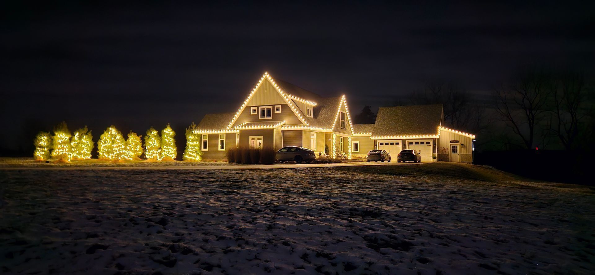 A snow-covered house lit with warm holiday lights at night, with glowing trees along the left side.