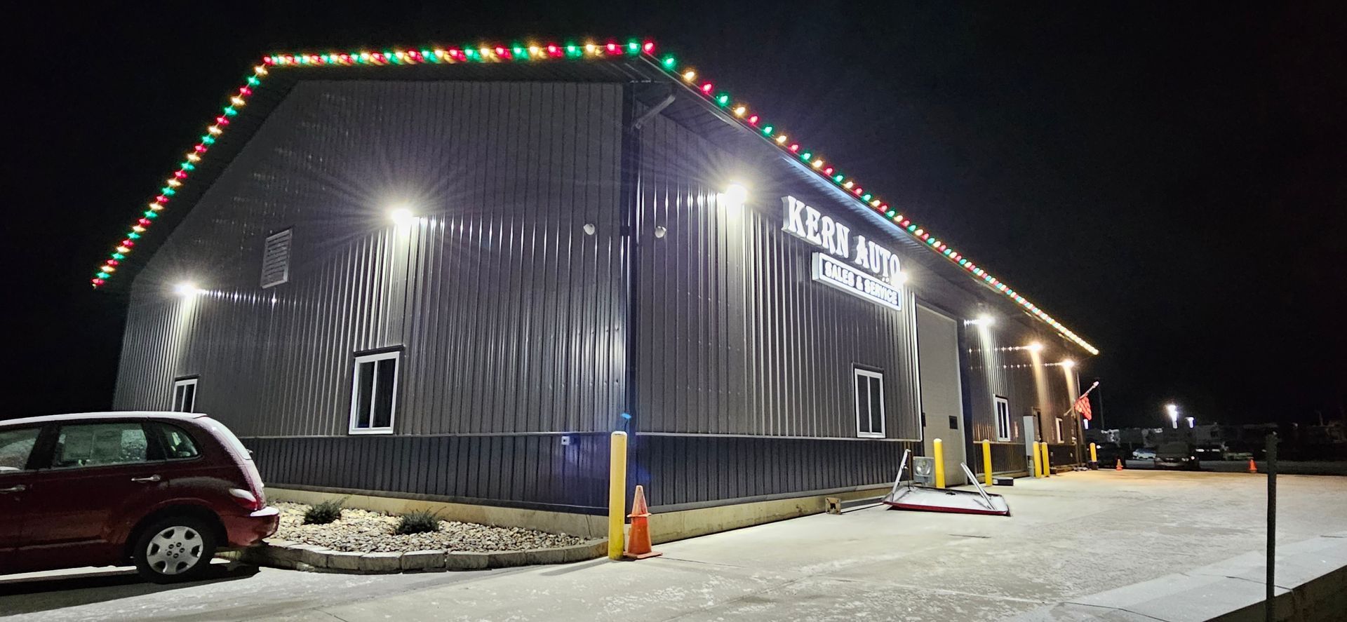 Snowy night outside a corrugated metal building with holiday lights and parked cars near the entrance
