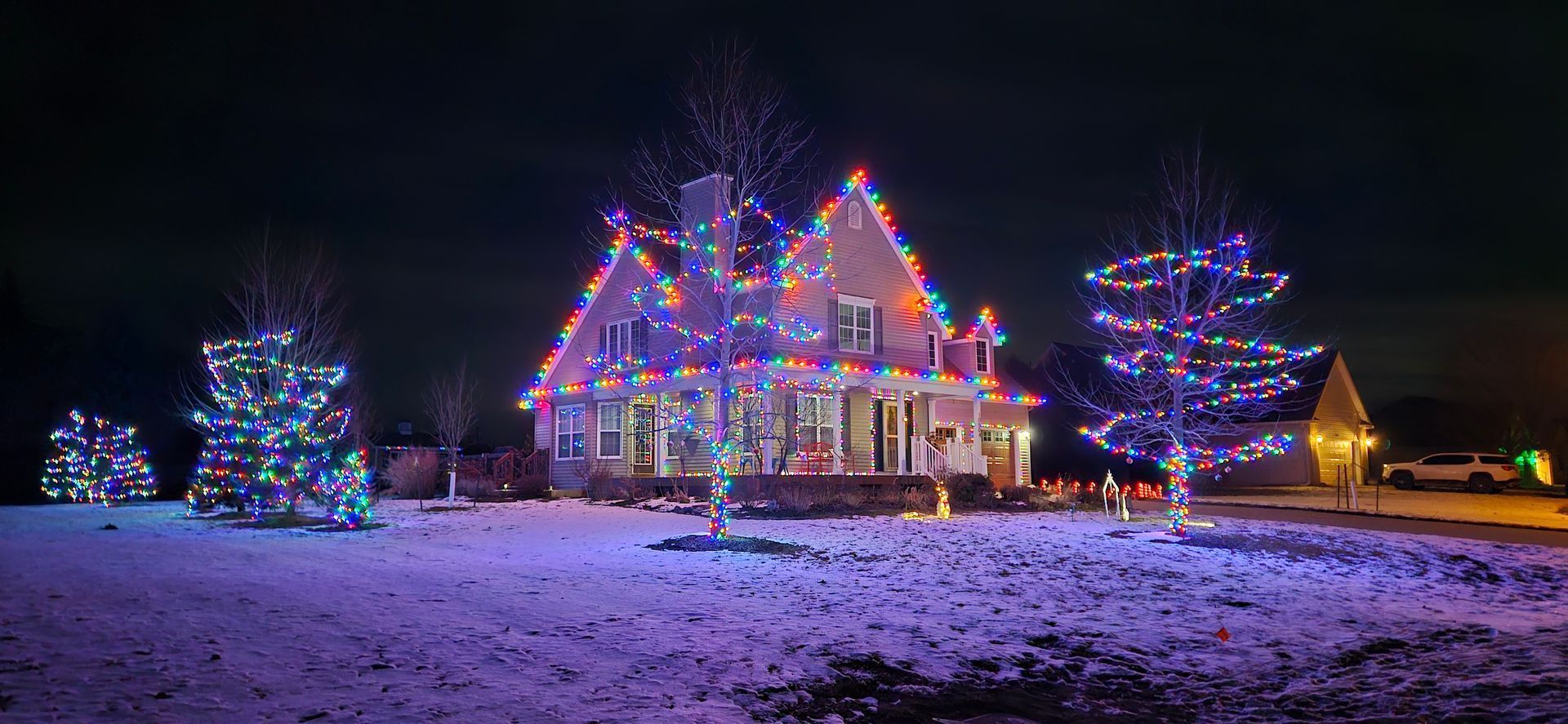 House and trees lit with colorful Christmas lights in a snowy yard at night