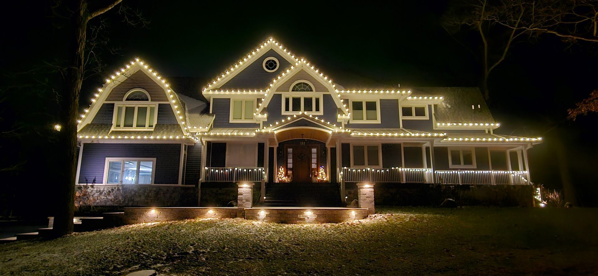 Large house outlined in warm white holiday lights at night, with a lit porch and lawn