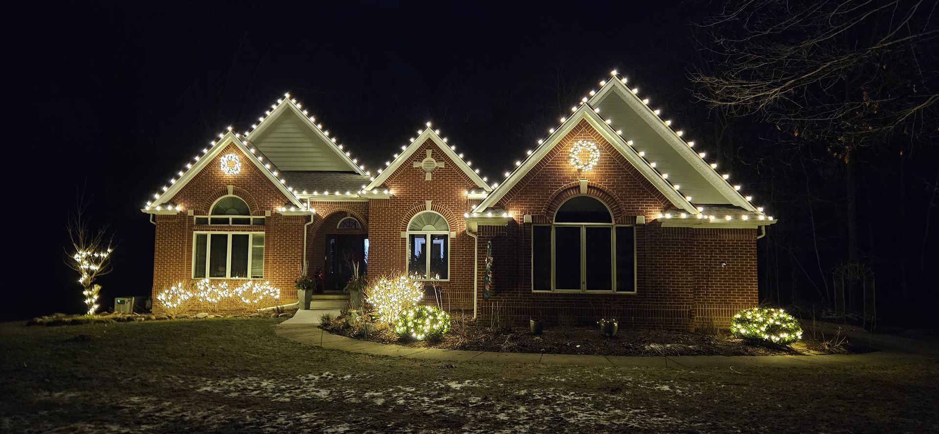 Brick house at night outlined with warm white holiday lights, surrounded by darkness and a snowy yard