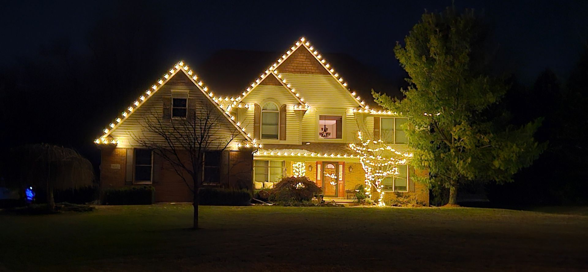 House at night outlined with warm white string lights, glowing windows, and trees in the yard