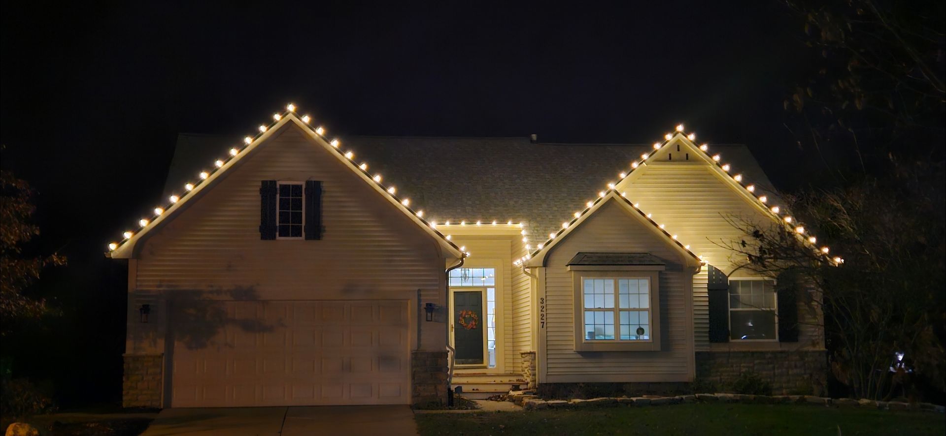 House at night outlined with warm white Christmas lights, front door glowing, garage on the left.