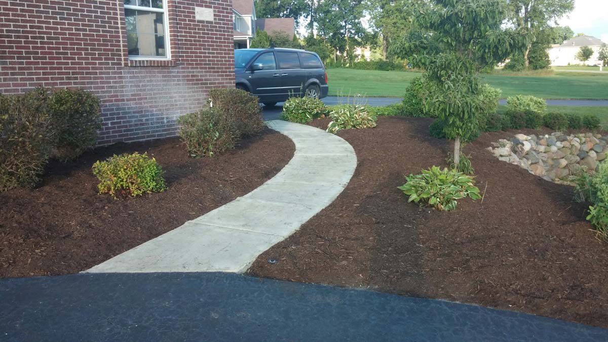 Front yard landscaped with a curved concrete path, mulched beds, shrubs, and a parked car near a brick house.