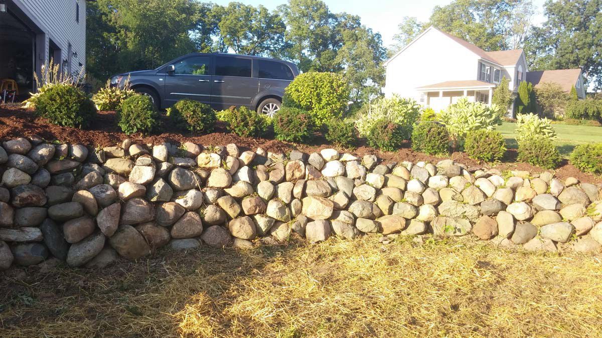 Stone retaining wall with shrubs in a yard, black car parked above it, and a house in the background.