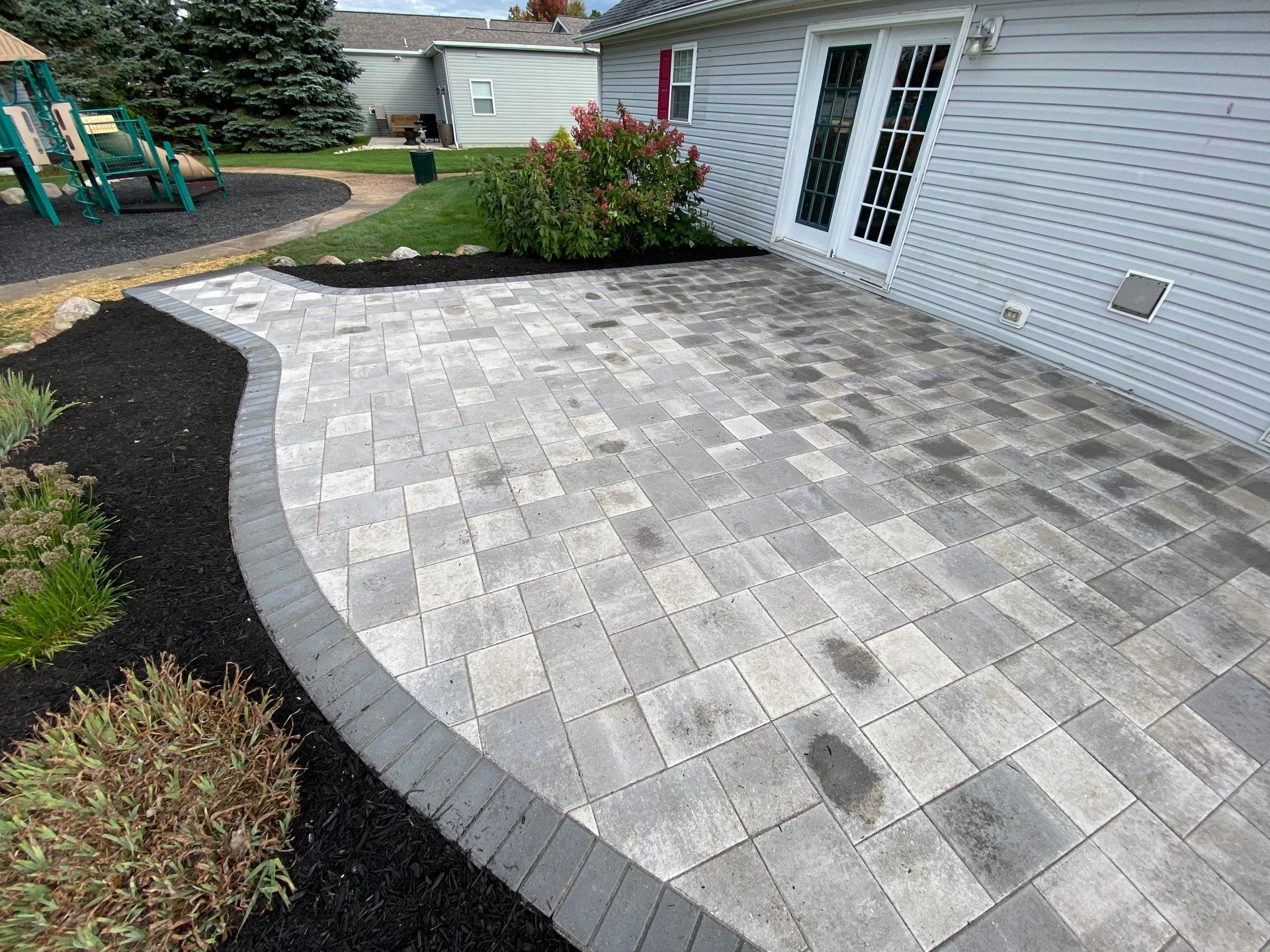 Gray paver patio beside a white house, bordered by curved edging and landscaped garden beds