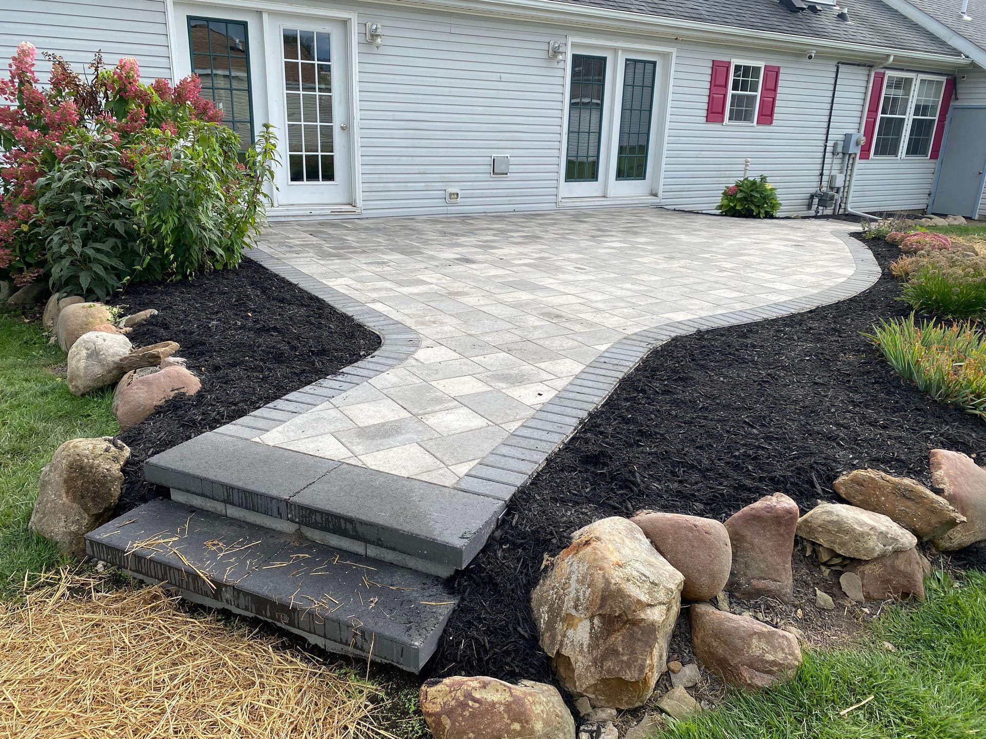 Stone paver walkway leading to a house entrance, bordered by dark mulch and rocks, with steps in front.