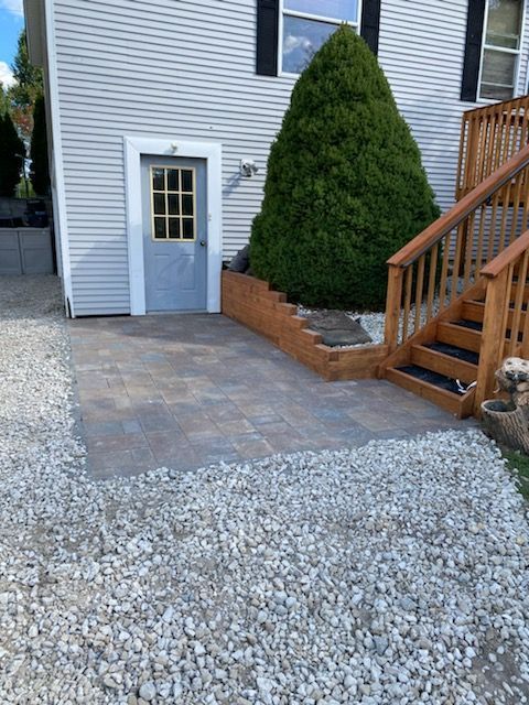 Backyard patio with stone pavers, gravel, a small blue door, and wooden stairs beside a house.