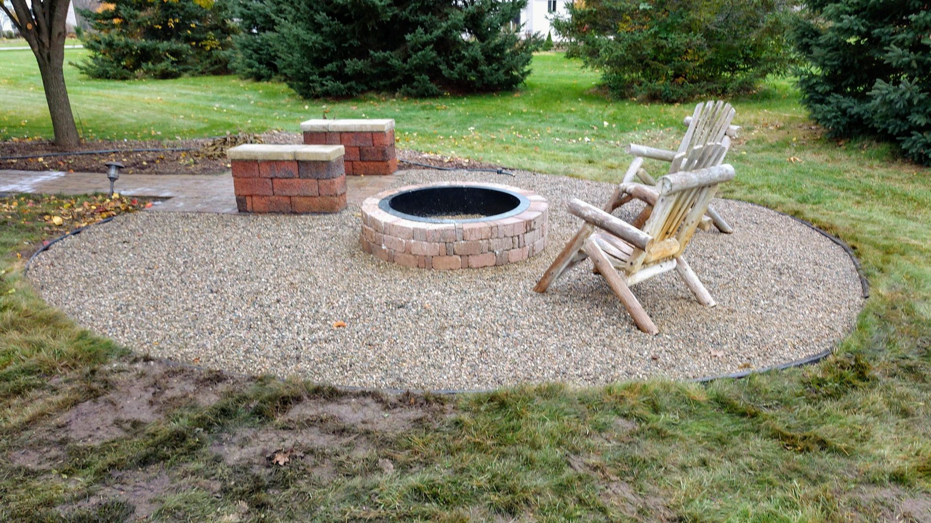 Outdoor fire pit area with gravel seating circle, brick bench, and wooden chairs in a grassy yard
