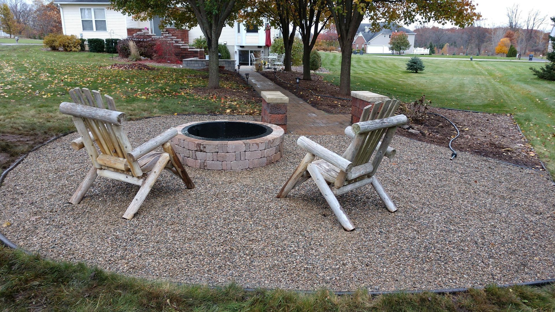 Backyard fire pit area with two wooden chairs on gravel and a brick ring, beside a lawn and steps.