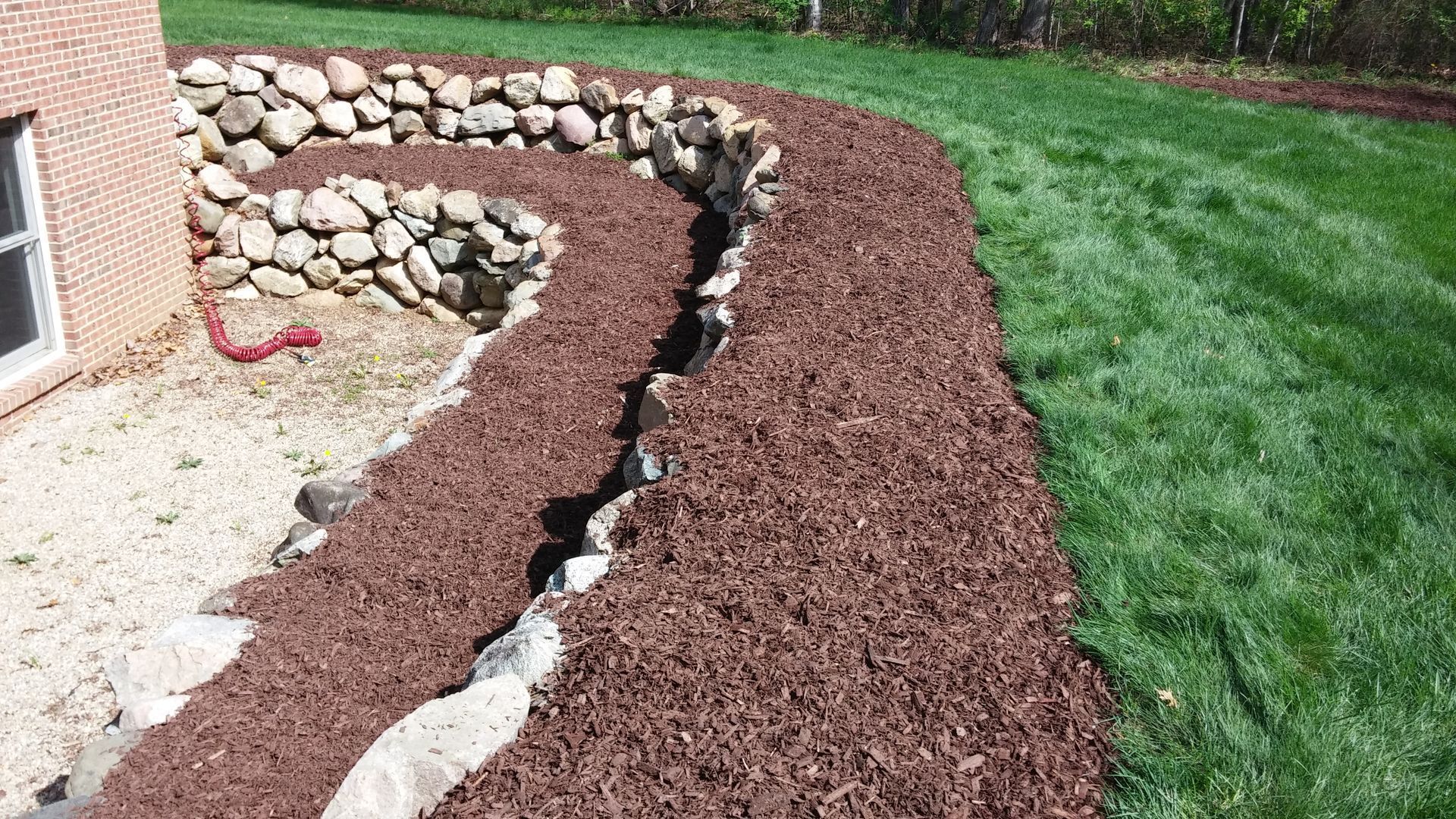 Curved mulch bed with stacked stone border beside a brick house and green lawn