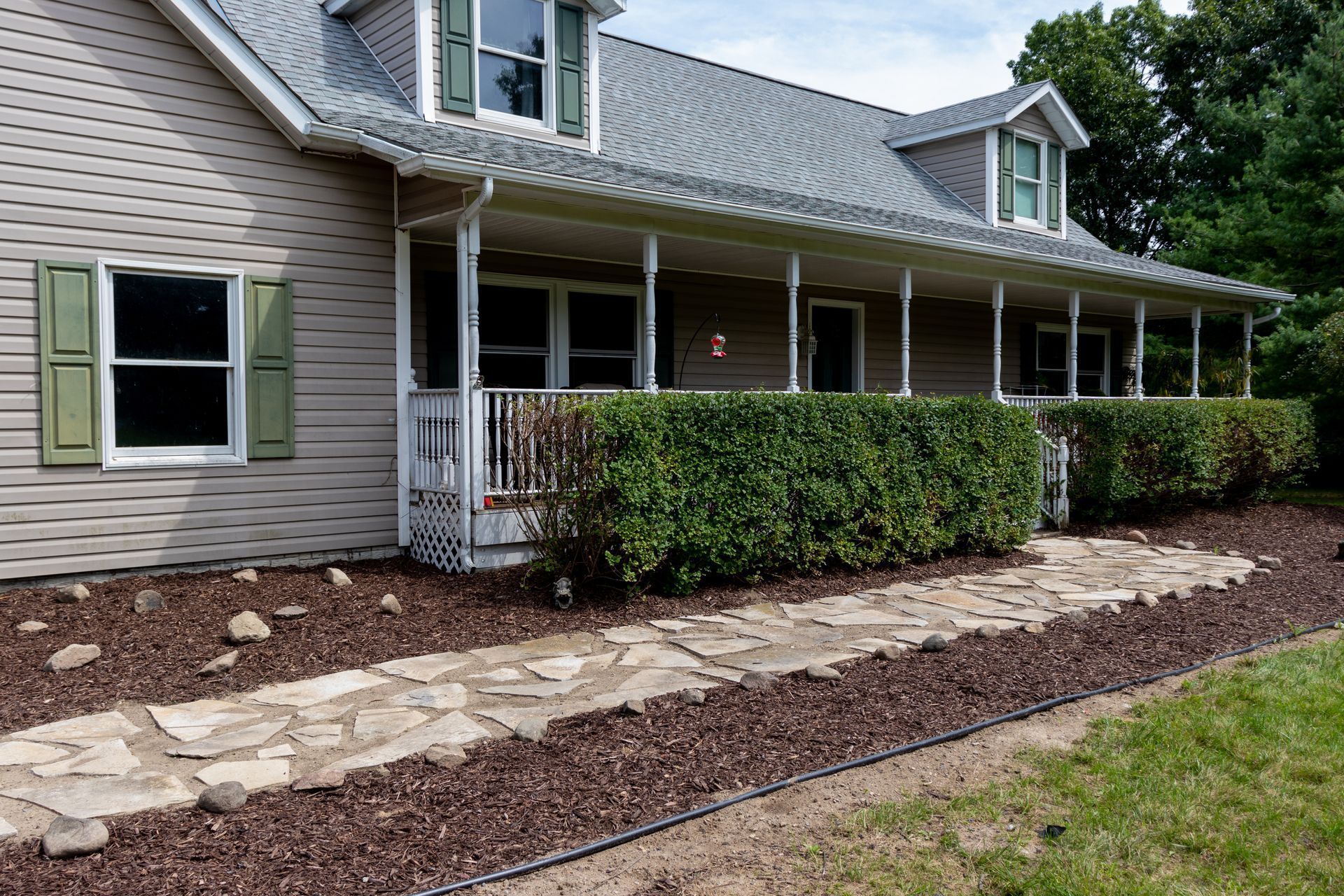 Gray house with a covered porch, green shutters, and a stone walkway bordered by mulch and hedges