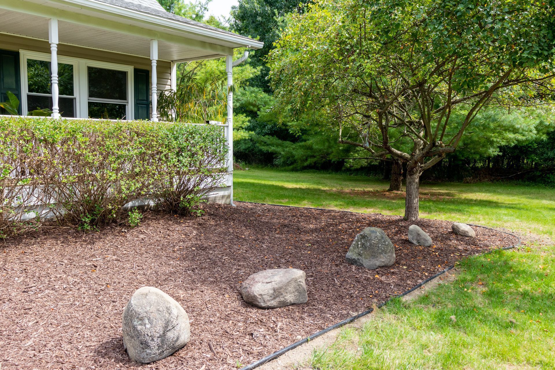 Front yard garden with rocks, mulch, and a small tree beside a house porch
