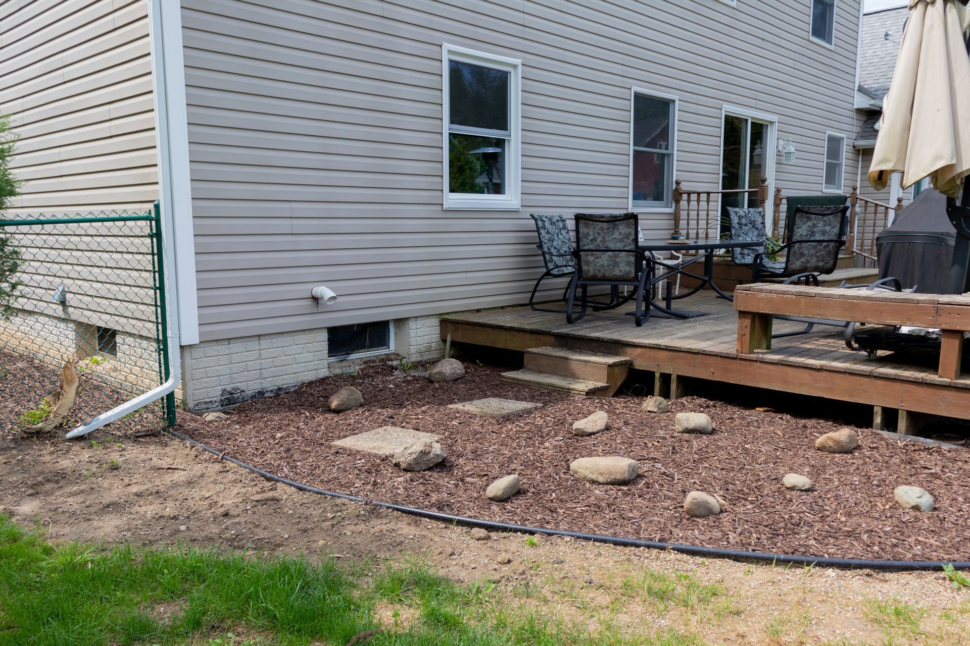 Backyard patio beside beige house with wooden deck, chairs, umbrella, and rock landscaping