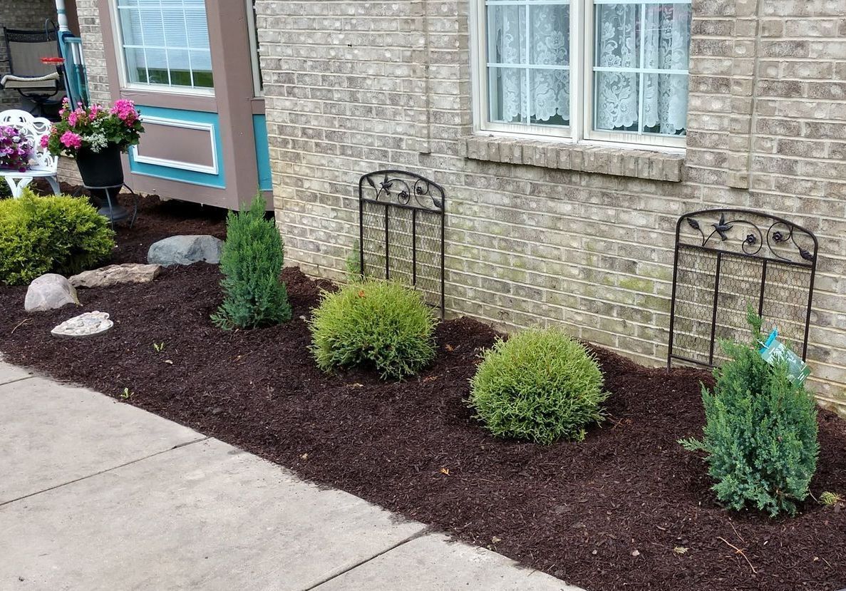 Landscaped flower bed with shrubs, mulch, and potted flowers beside a brick house and sidewalk