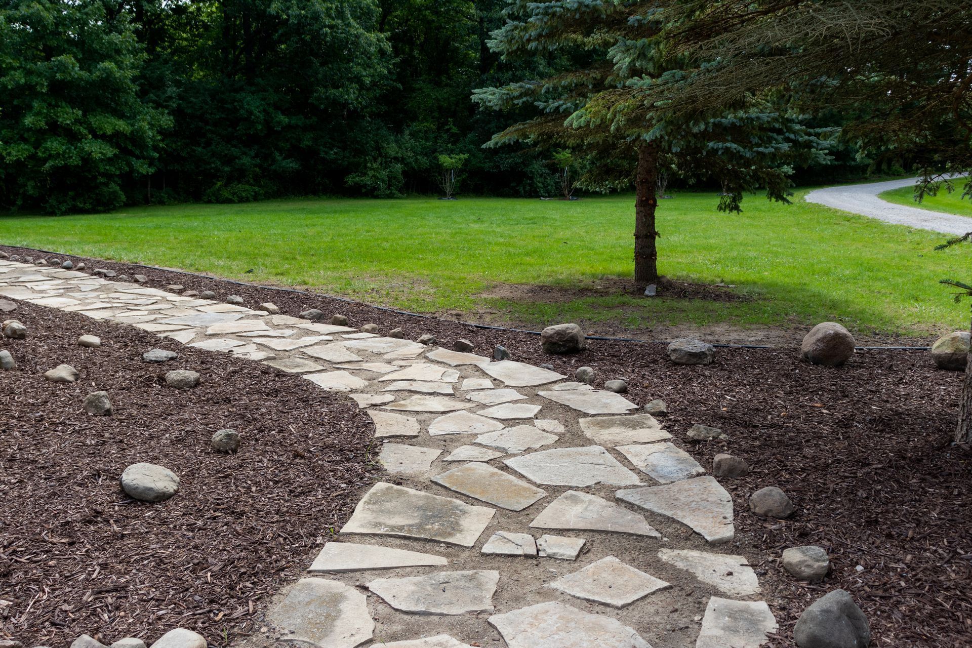 Stone garden path curving through mulch toward a grassy lawn with trees