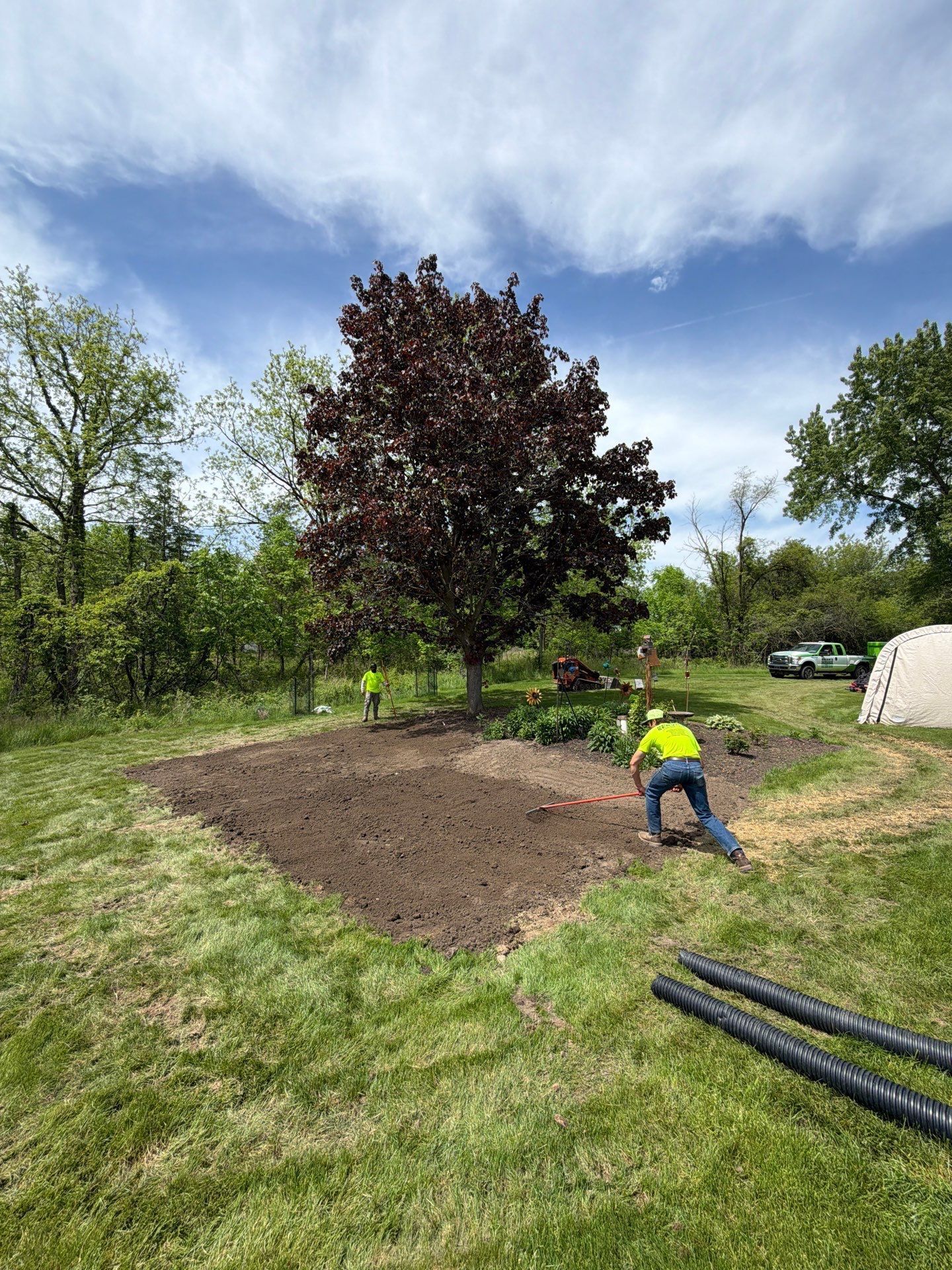 Person tilling a large dirt patch in a grassy backyard under a partly cloudy sky