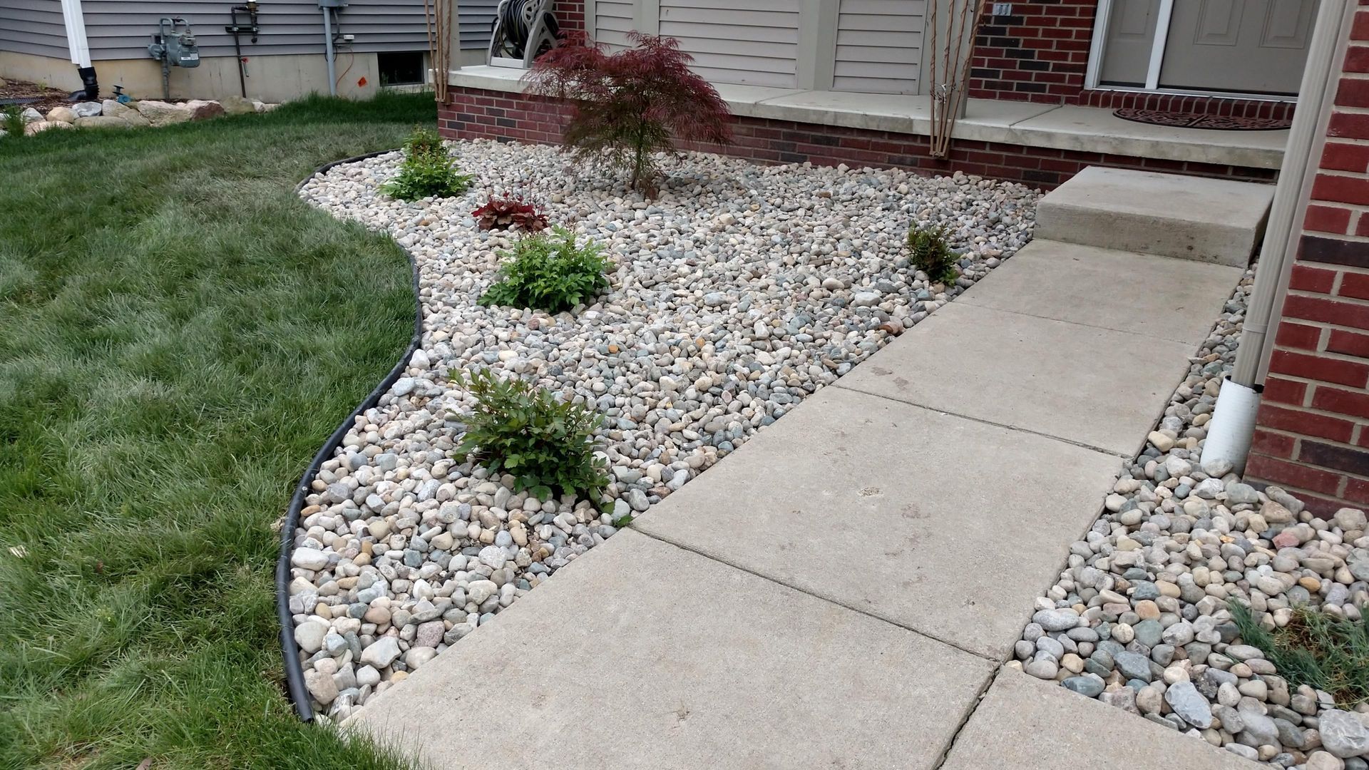 Curved concrete walkway beside a white rock garden and house front porch with small shrubs and grass