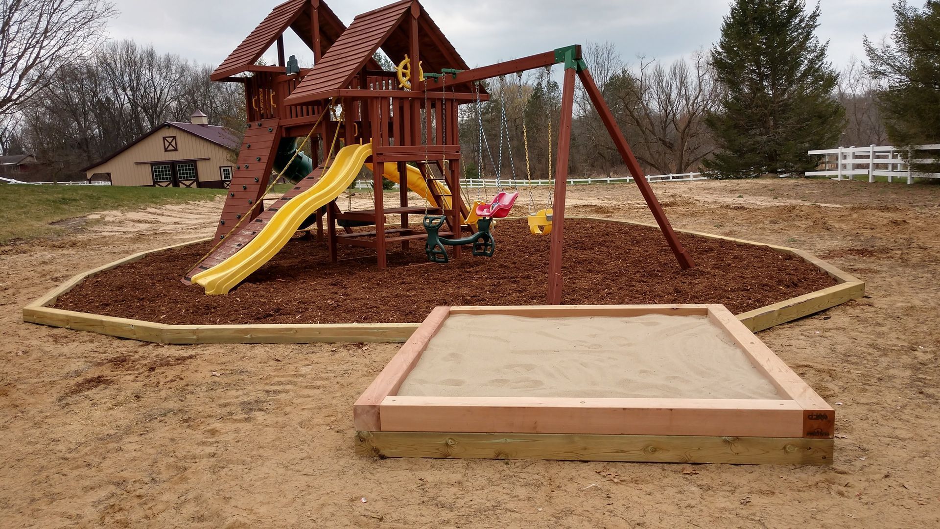 Backyard playground with wooden playset, yellow slide, and sandbox in front on wood chips.