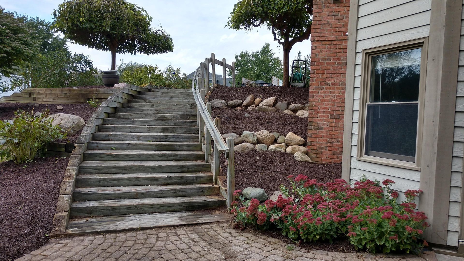 Outdoor stone staircase beside a house, with landscaped rocks, flowers, and a tree at the top