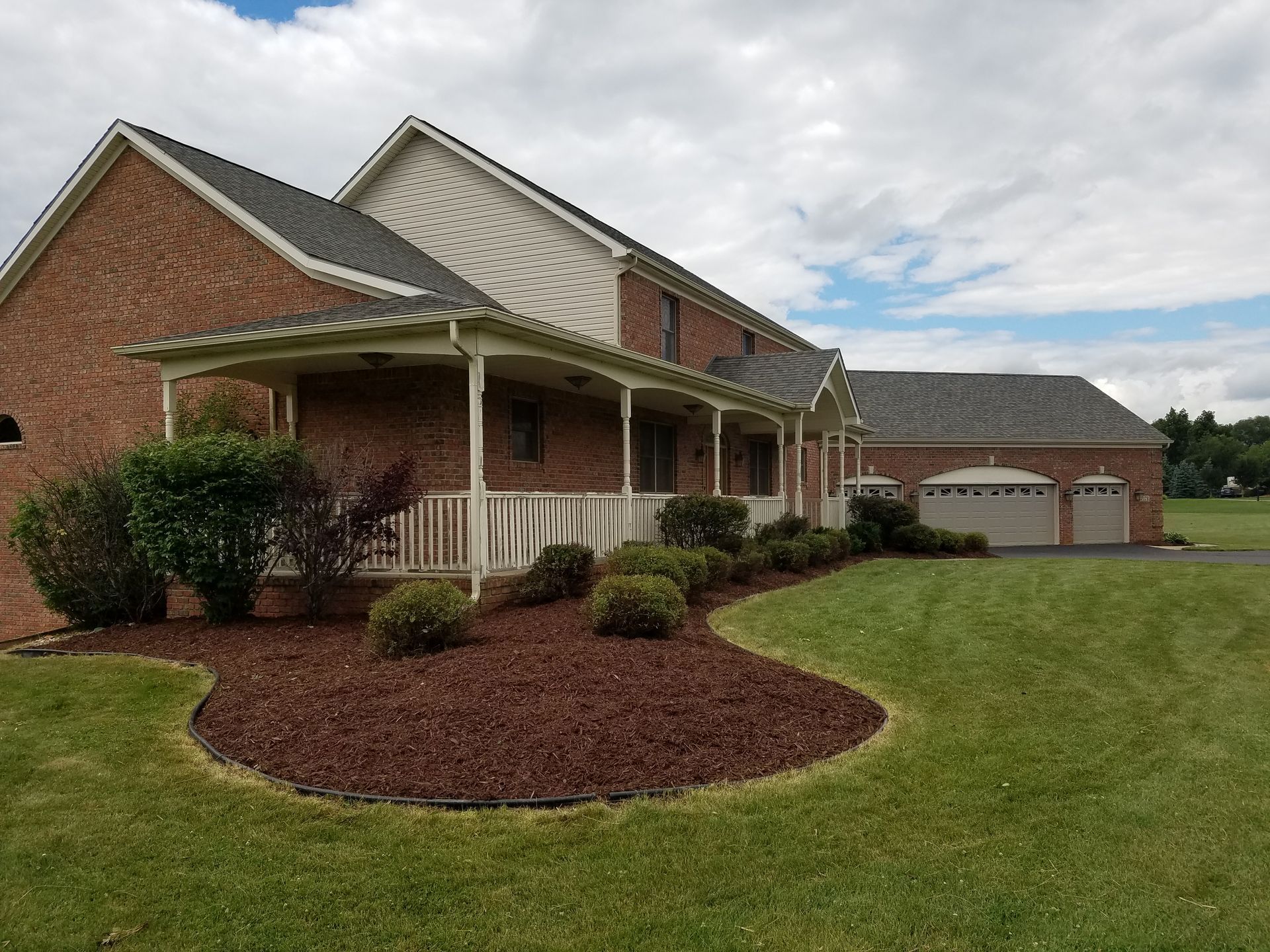 Brick house with covered porch, landscaped front yard, and detached garage under a cloudy sky