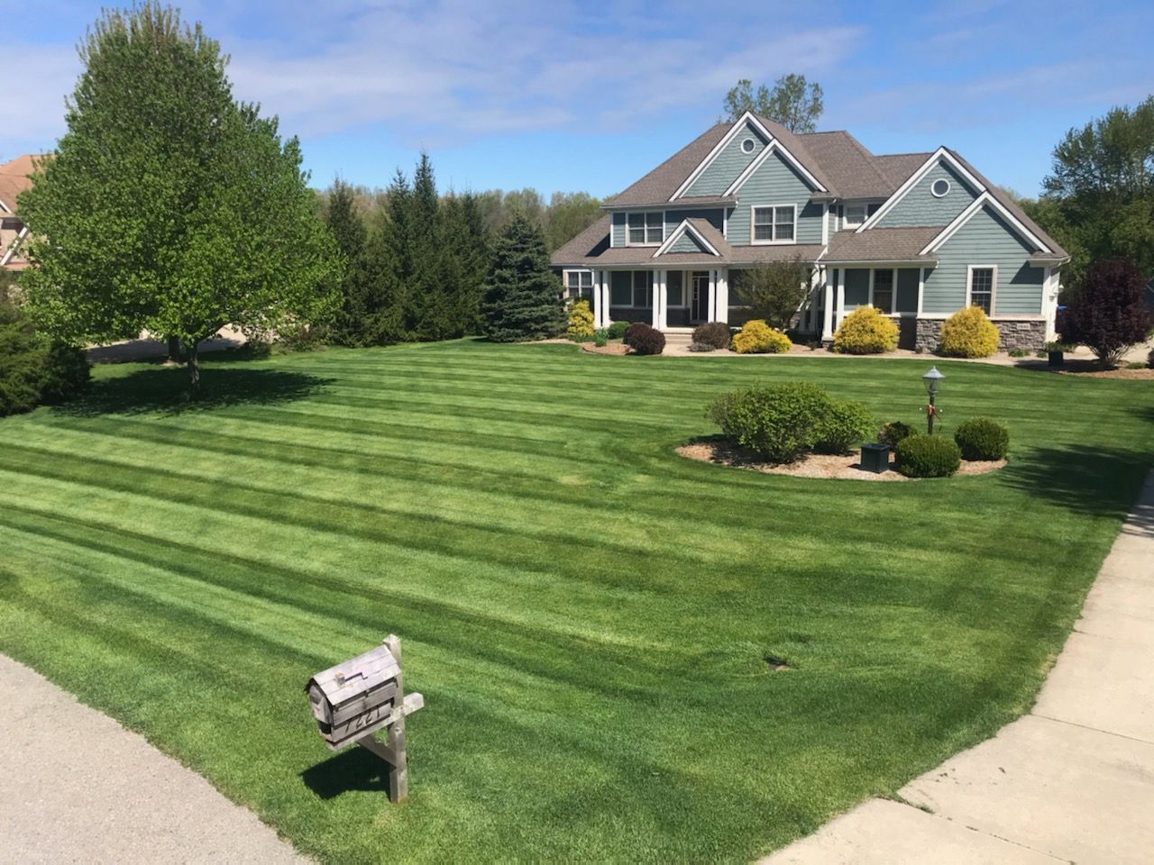 Suburban house with manicured striped lawn, shrubs, and a mailbox under a blue sky
