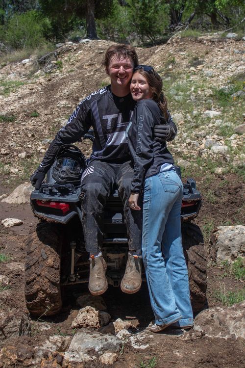 Two people sitting on a muddy ATV outdoors, smiling and posing together