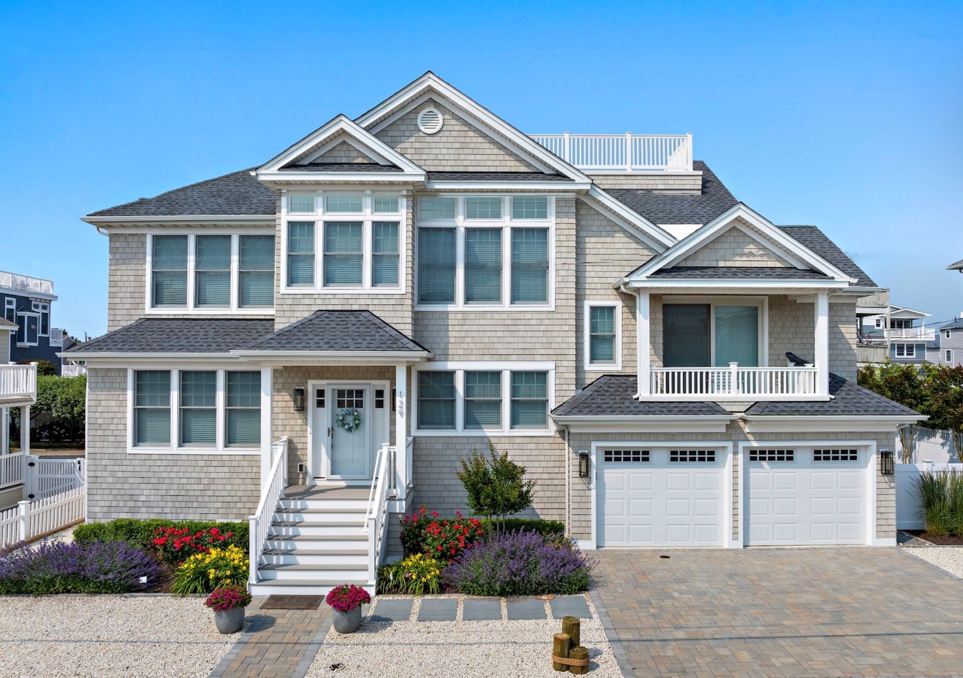 Two-story beach house with gray shingles, white trim, white garage doors, and colorful landscaping under a blue sky.
