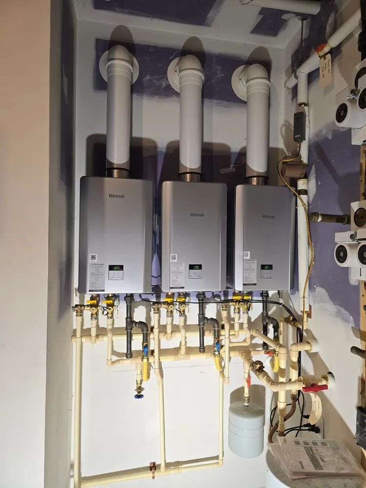 Three silver tankless water heaters mounted on a wall in a mechanical room with interconnected white piping.