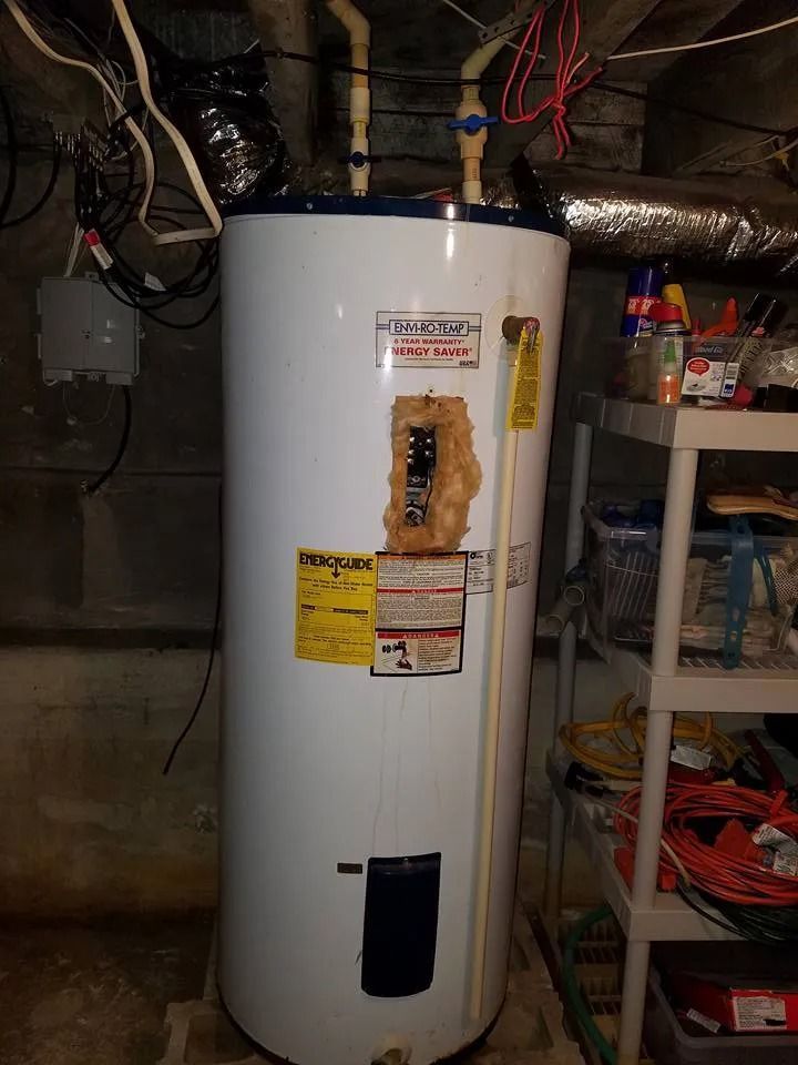 A white electric water heater stands in a basement next to storage shelving, with exposed wiring and pipes above it.