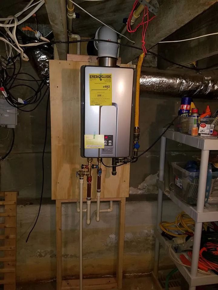 A tankless water heater mounted on a wooden board in a basement, with visible plumbing connections and exhaust vent.