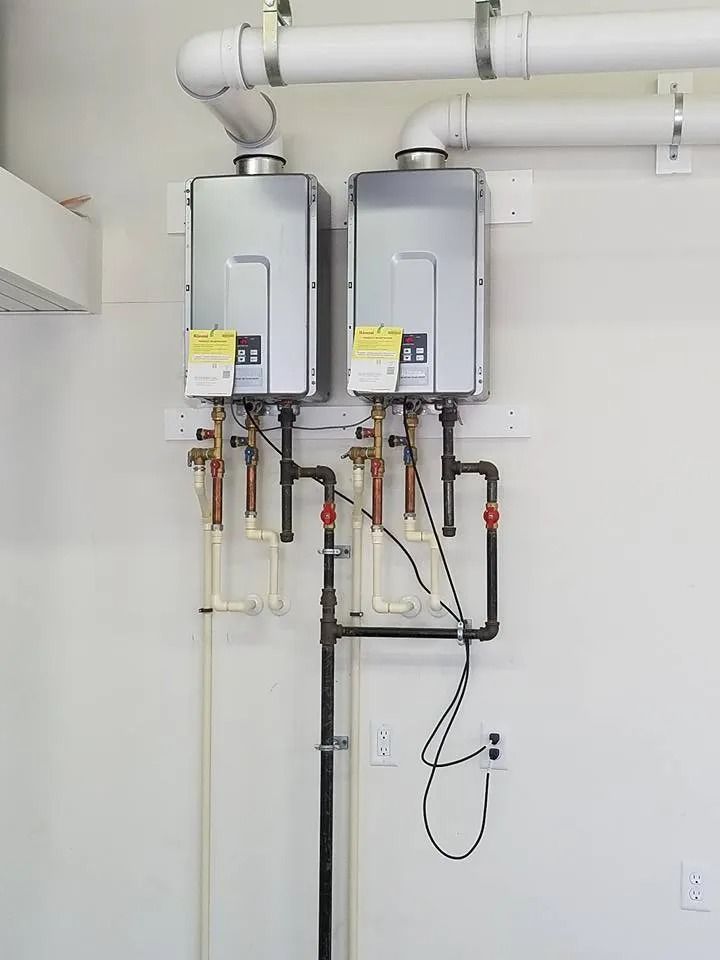 Two wall-mounted tankless gas water heaters connected by white exhaust pipes and black gas lines against a white wall.