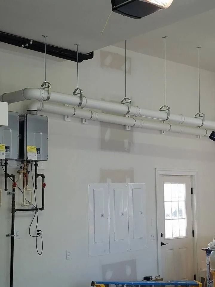 Two tankless water heaters mounted on a wall with white vent pipes suspended from the ceiling in a garage.