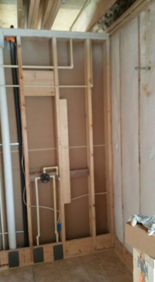 Framing for a wall showing exposed wooden studs, plumbing pipes, and electrical wiring in an unfinished interior room.