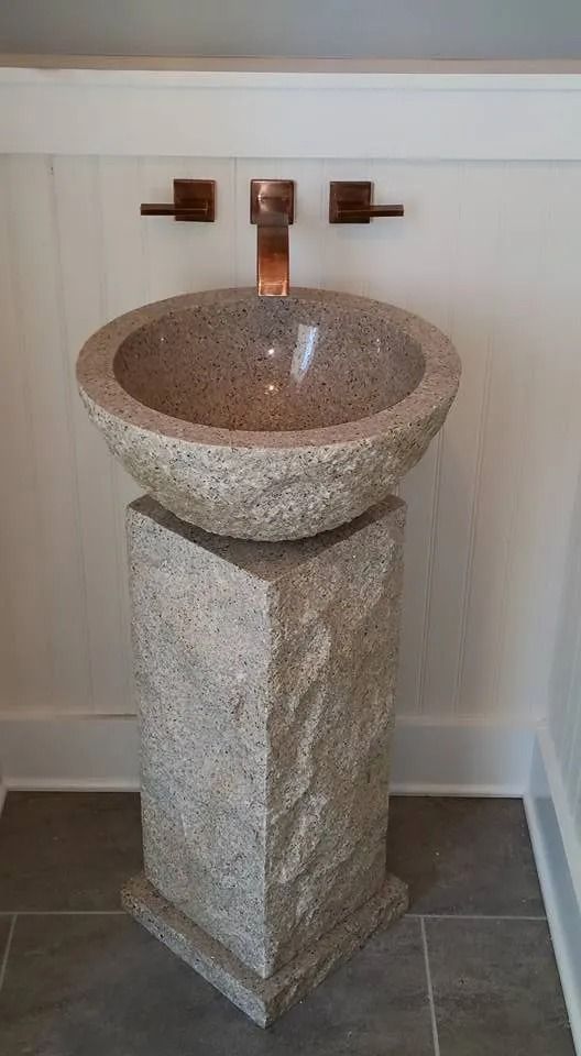 A stone pedestal sink with a matching bowl-shaped basin and a copper wall-mounted faucet, set against a white wall.