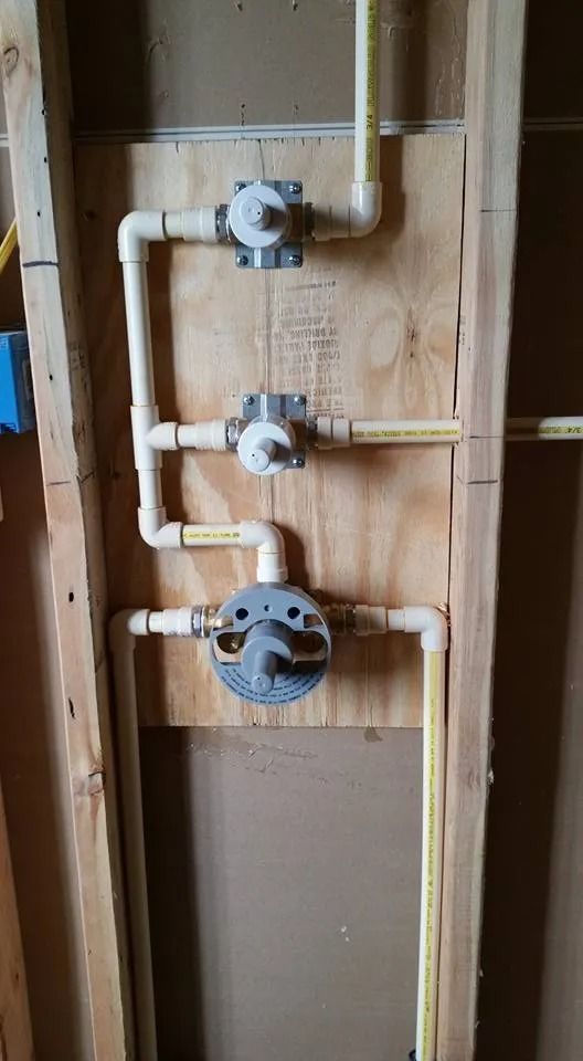 A shower plumbing assembly with three valve controls connected by light-colored CPVC pipes on a wooden wall panel.