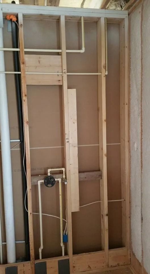 An interior stud wall under construction showing exposed wooden framing, beige backing panels, and plumbing pipes.