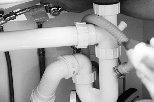 Close-up of hands installing white plastic plumbing pipes and a flexible drainage hose under a sink.