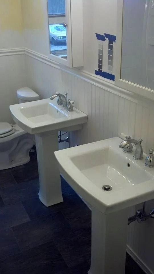A bathroom features two white pedestal sinks, a white toilet, dark flooring, and paint swatches taped to the wall.